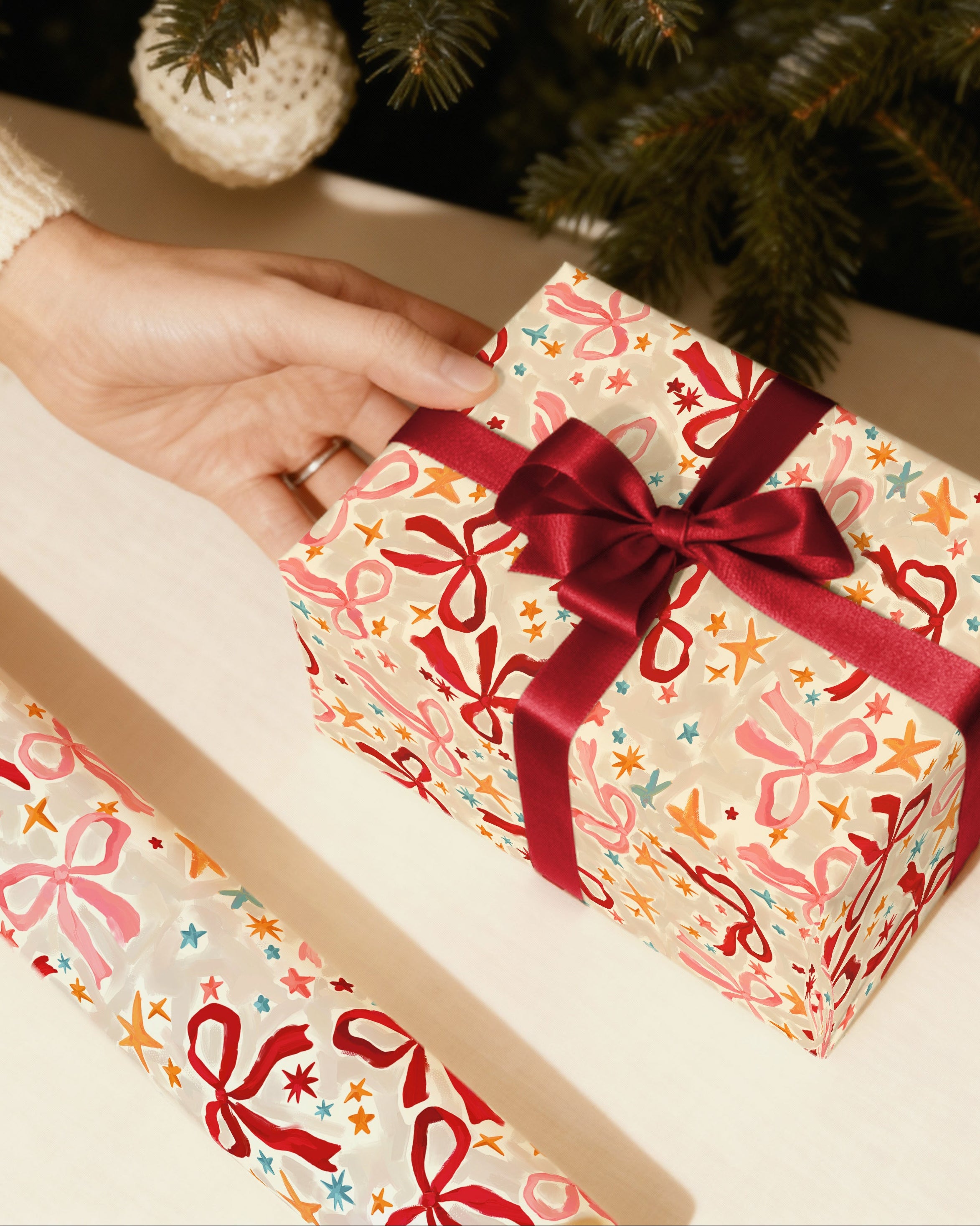 Person opening a gift wrapped in colorful paper with a red ribbon near a Christmas tree.