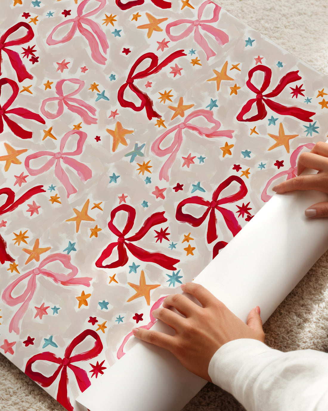 Person unrolling a colorful gift wrap with red bows on a beige carpet