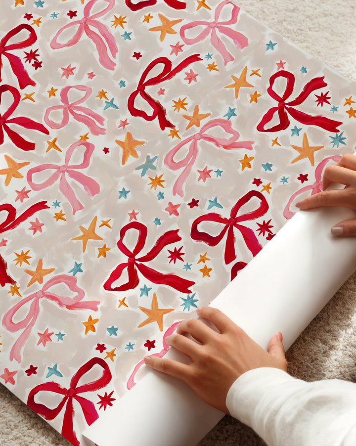 Person unrolling a colorful gift wrap with red bows on a beige carpet
