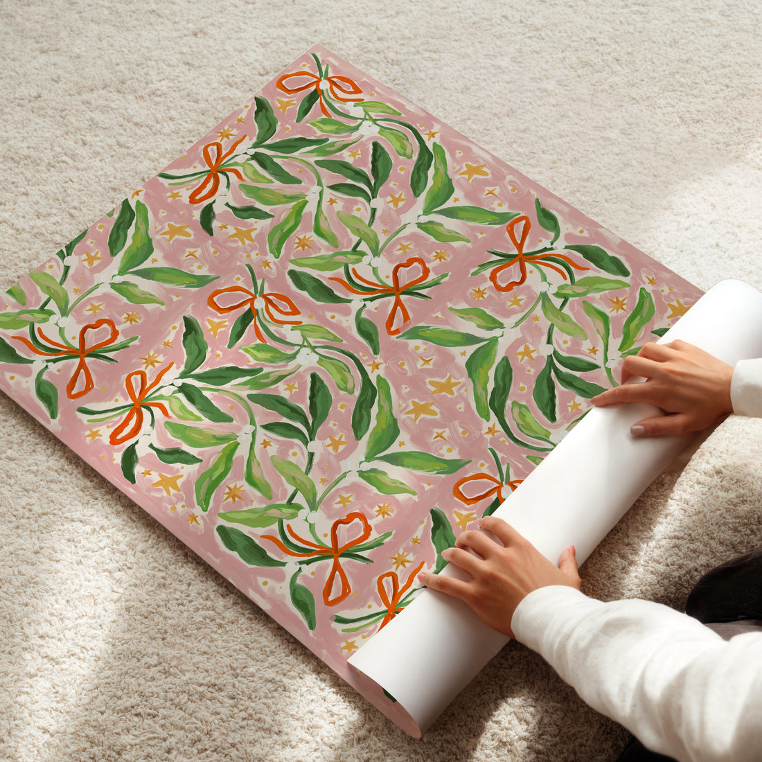 Person unrolling a roll of pink wrapping paper with green mistletoe and orange ribbons on a light-colored floor.