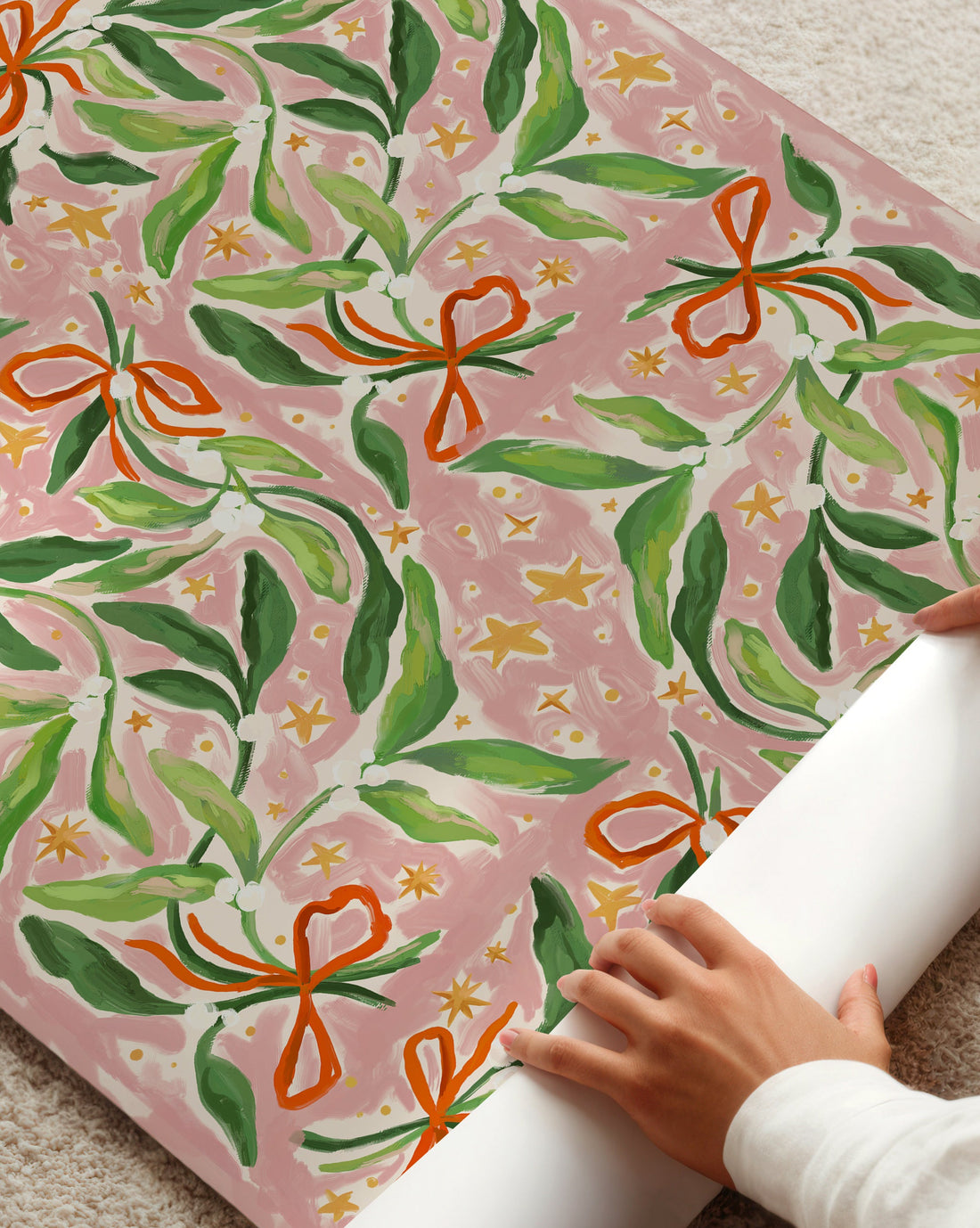 Person unrolling a roll of pink wrapping paper with green mistletoe and orange ribbons on a light-colored floor.